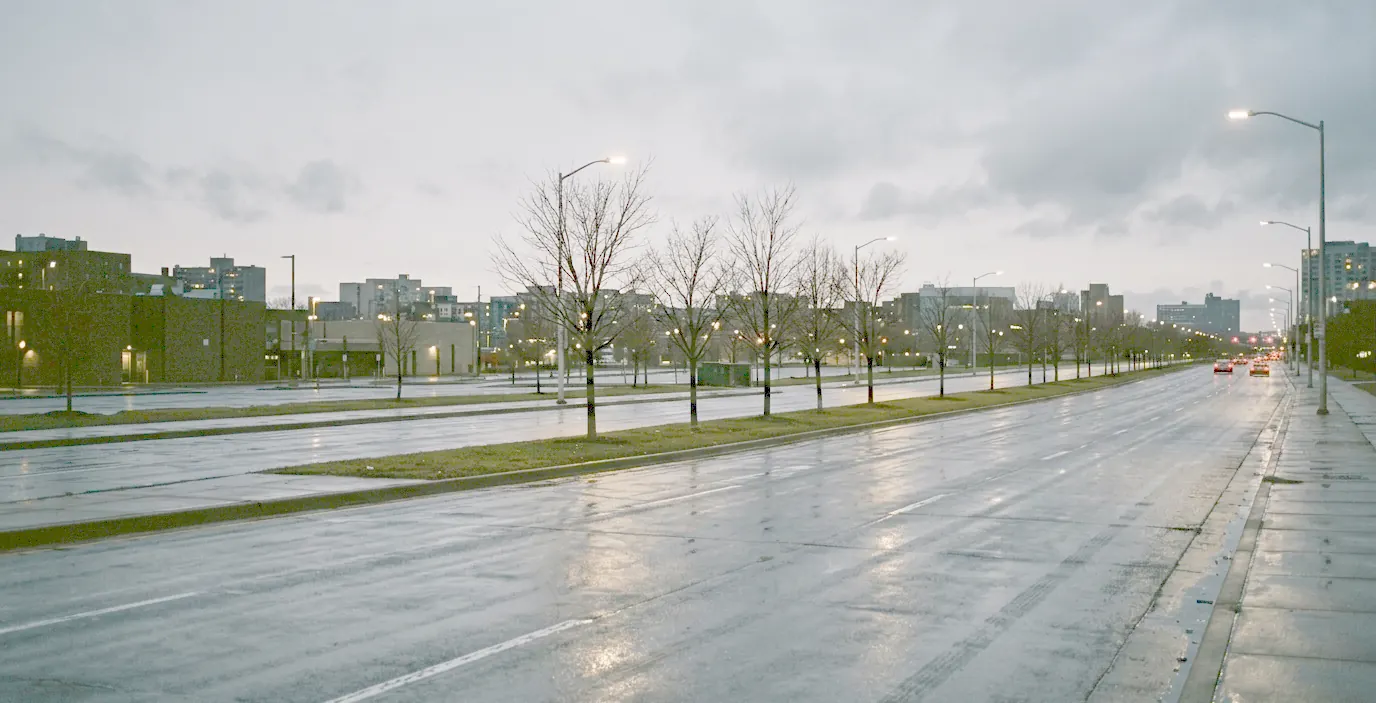 A wide city boulevard at dusk after rain, wet asphalt reflecting the street lights