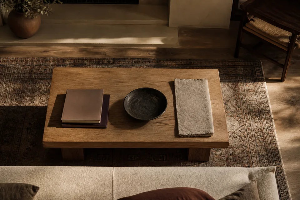 A low oak table seen from above with books, a dark bowl and a folded linen runner.