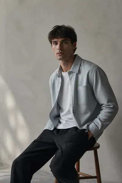 Model seated on a low stool in a pale grey overshirt beside a plaster wall