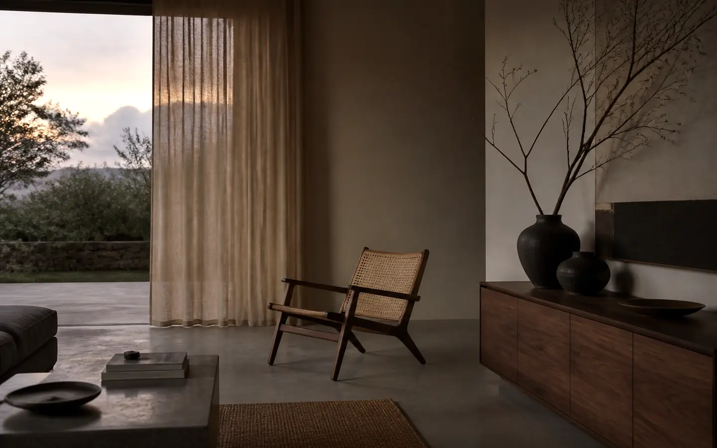 Dim living room at dusk with a linen curtain, a cane lounge chair and a walnut sideboard