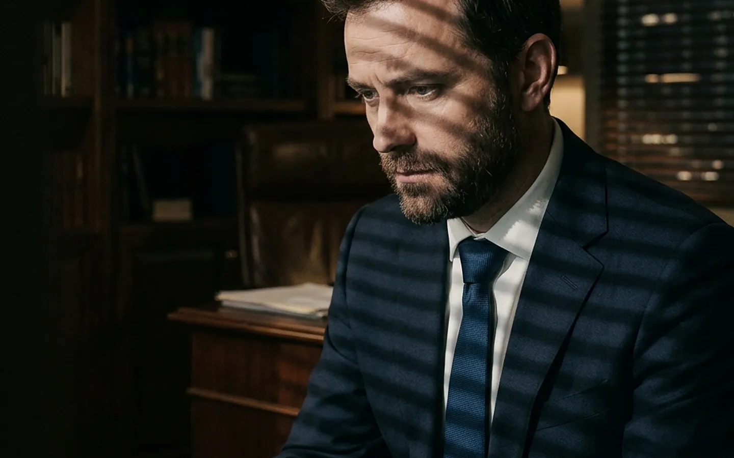 A lawyer in a dark suit reading a document, venetian blind shadows across his face