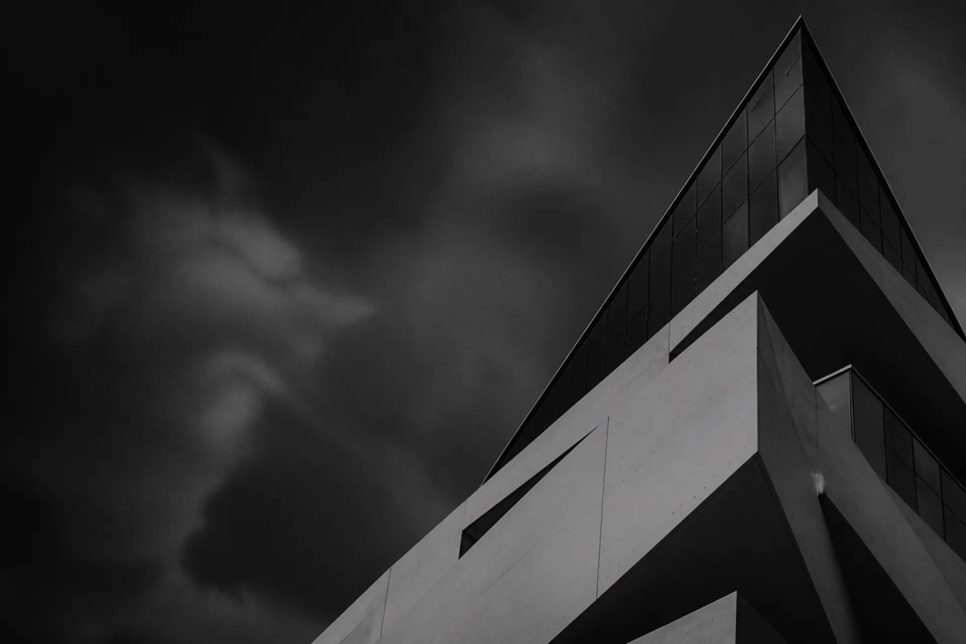 Black and white view up at an angular concrete and glass building under a stormy sky