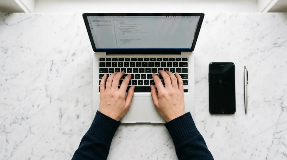 Hands typing on a silver laptop beside a phone and pen on a marble desk