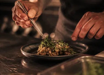 A cook finishing a plate of greens at the kitchen pass