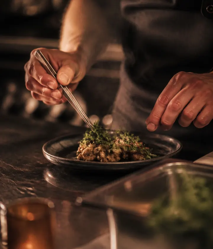 A cook plating greens with tweezers at the pass